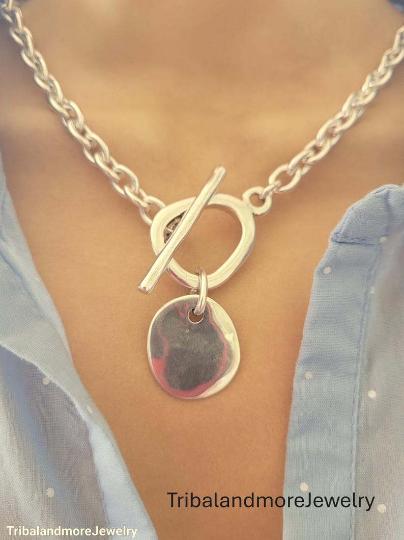 May include: A silver-toned necklace with a large link chain and a toggle clasp. The pendant is a small, round, hammered disc. The necklace is worn against a light blue shirt. The text "TribalandmoreJewelry" is at the bottom.