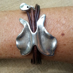 May include: A silver butterfly charm bracelet with brown leather straps. The charm is a stylized butterfly with a smooth, rounded surface.