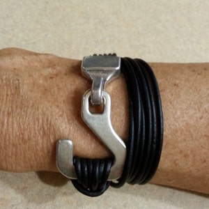 May include: A black leather bracelet with a silver anchor-shaped clasp. The bracelet is made of multiple strands of leather and has a simple, modern design.