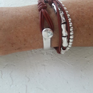 May include: A brown leather bracelet with silver beads and a silver metal charm. The bracelet is wrapped around a wrist.
