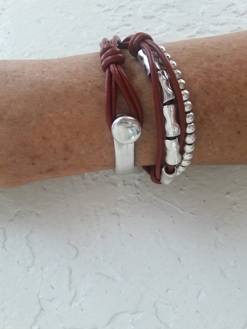 May include: A brown leather bracelet with silver beads and a silver metal charm. The bracelet is wrapped around a wrist.