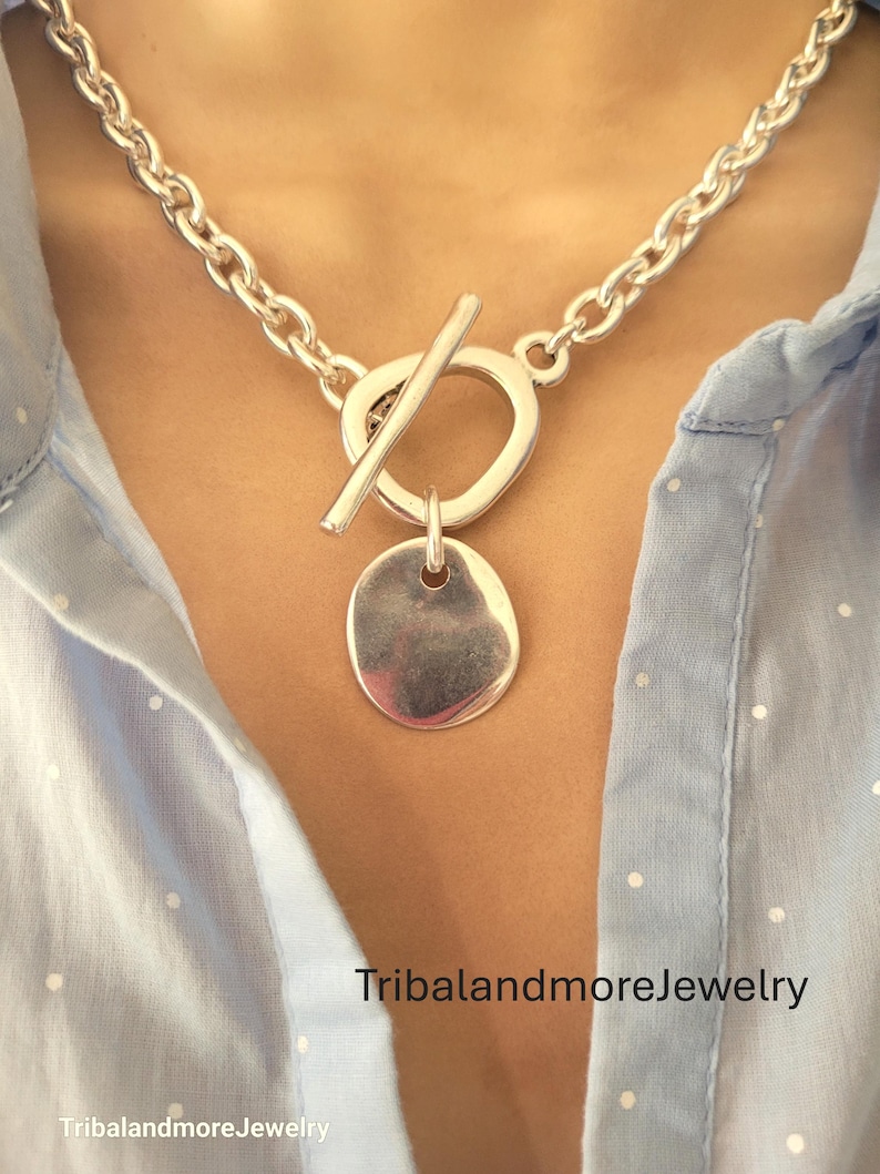 May include: A silver-toned necklace with a toggle clasp and a round pendant. The chain is a chunky link design. The necklace is worn over a light blue shirt. The text "TribalandmoreJewelry" is visible.