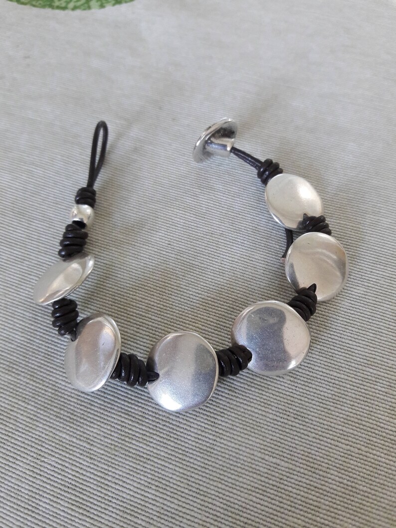 Puede incluir: Un brazalete de cuero negro con cuentas redondas plateadas. El brazalete tiene un cierre de bot&oacute;n.