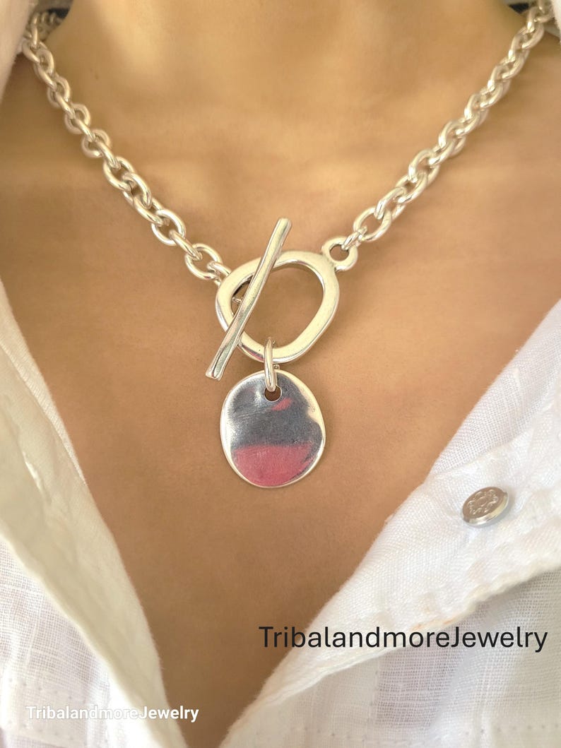 May include: A silver-toned necklace with a toggle clasp and a pendant. The pendant is an oval shape with a polished finish. The necklace has a chunky chain design. The text "TribalandmoreJewelry" is visible at the bottom of the image.