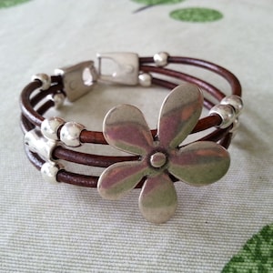 May include: A brown leather bracelet with three strands and a silver flower charm. The bracelet has silver beads spaced along the strands.