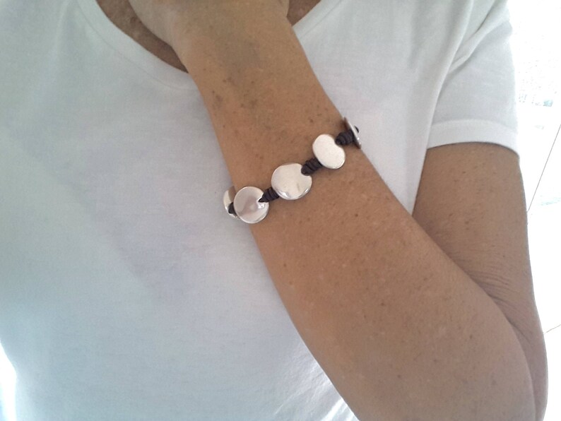 Puede incluir: Un brazalete de plata con abalorios redondos conectados por un cord&oacute;n marr&oacute;n.