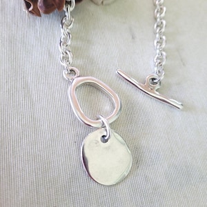 May include: A silver-toned bracelet with a toggle clasp and a chain link design. The bracelet features a large oval-shaped link and a small, round, flat charm. The jewelry is displayed on a light gray surface.