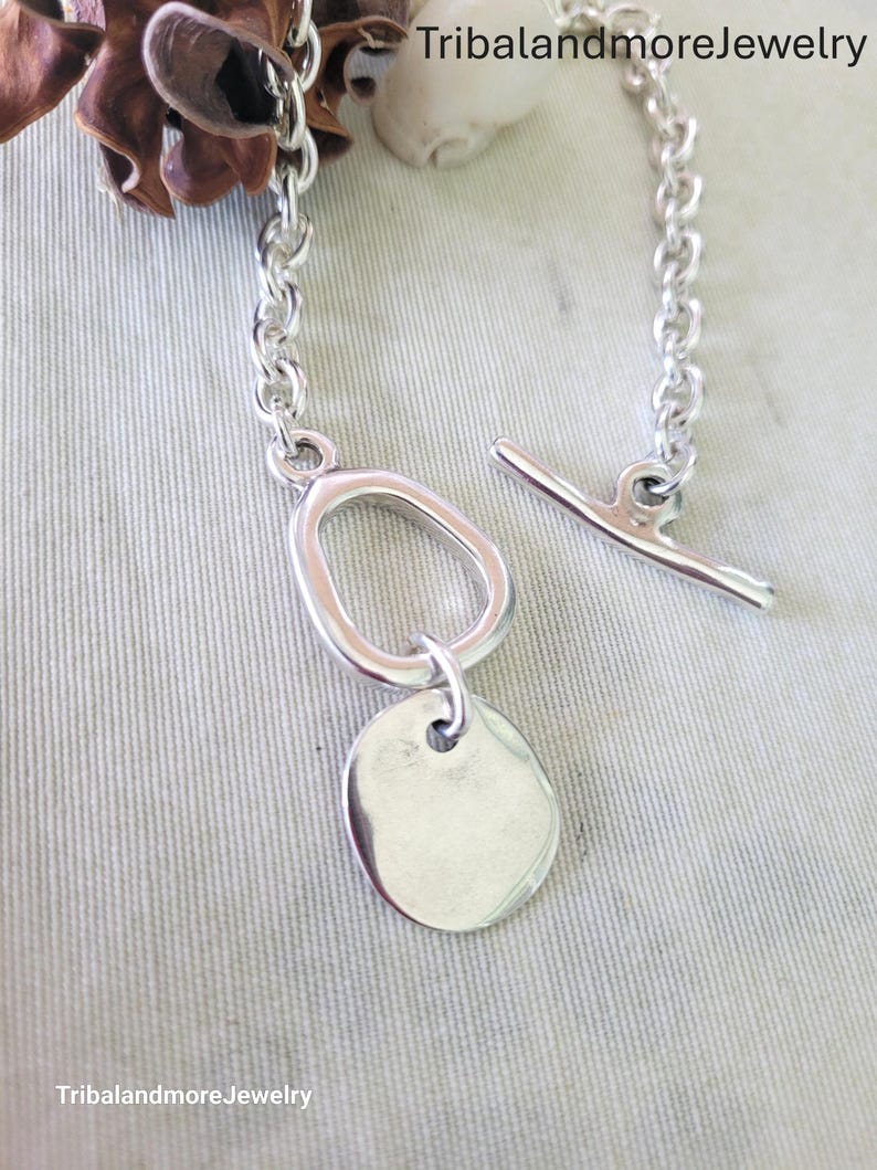 May include: A silver-toned bracelet with a toggle clasp and a chain link design. The bracelet features a large oval-shaped link and a small, round, flat charm. The jewelry is displayed on a light gray surface.