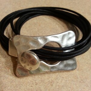 May include: A silver bracelet with a hammered metal design and a black leather band. The bracelet is made of multiple strands of leather.