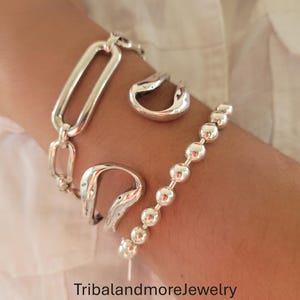 May include: A collection of silver-toned jewelry, including a bracelet with a chain of connected rectangles, a bracelet with small round beads, and two curved rings. The jewelry is displayed on a person's arm against a white background. The text "TribalandmoreJewelry" is visible.