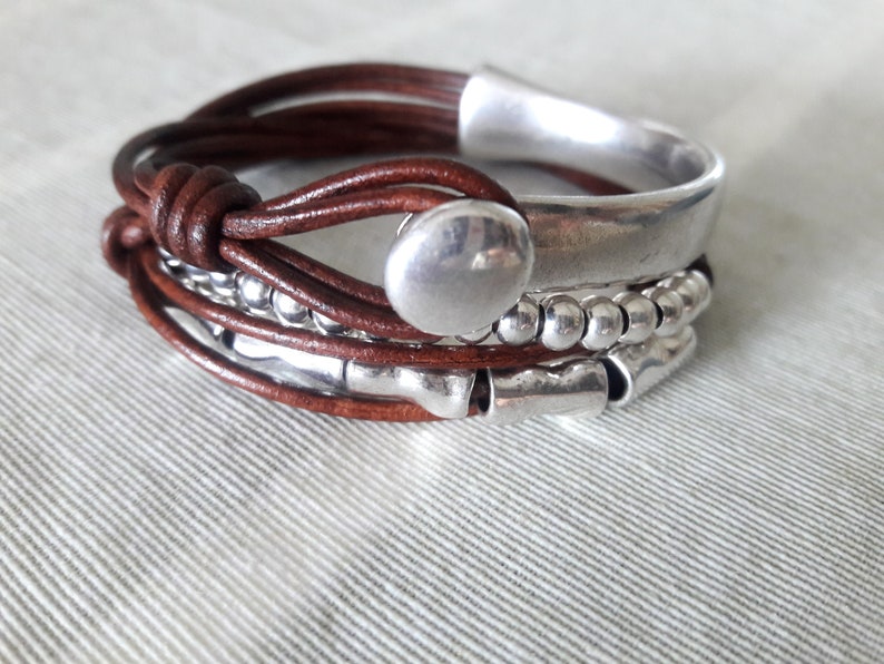 May include: A brown leather bracelet with silver beads and a large silver charm. The bracelet is layered with multiple strands of leather and metal.