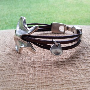 May include: A brown leather bracelet with a silver charm and a silver clasp. The charm is shaped like a bird.
