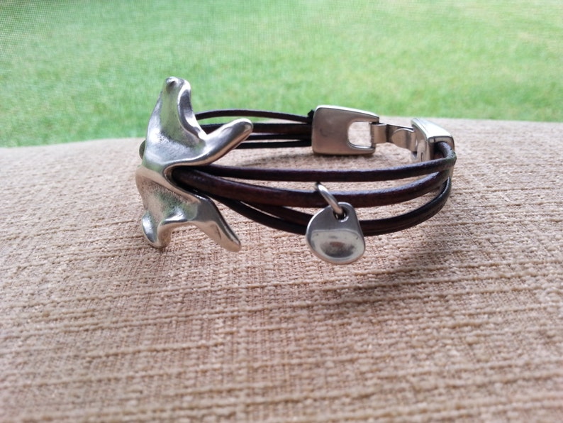 May include: A brown leather bracelet with a silver charm and a silver clasp. The charm is shaped like a bird.