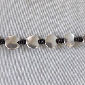 Puede incluir: Un brazalete de plata con abalorios redondos de metal conectados por un cord&oacute;n negro.