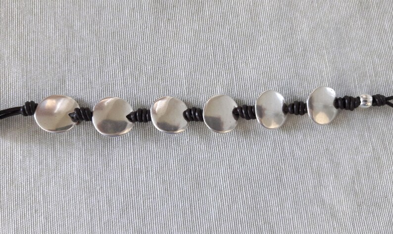 Puede incluir: Un brazalete de plata con abalorios redondos de metal conectados por un cord&oacute;n negro.