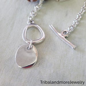 May include: A silver-toned bracelet with a toggle clasp. The bracelet features a chain link design, a circular pendant, and a unique asymmetrical ring. The text "TribalandmoreJewelry" is visible.