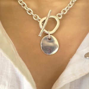 May include: A silver-toned necklace with a toggle clasp and a pendant. The necklace features a chunky chain and a circular toggle clasp. The pendant is a small, oval-shaped disc. The necklace is worn over a white shirt.