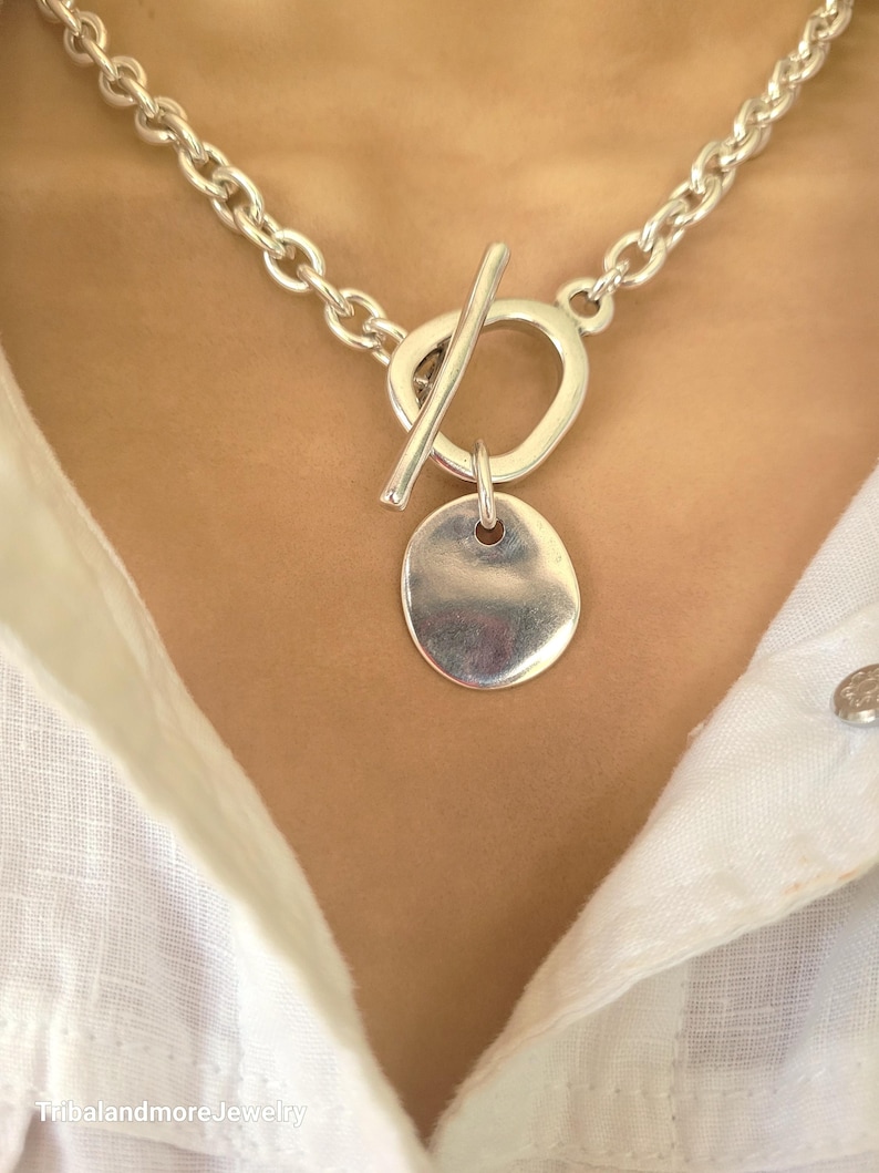 May include: A silver-toned necklace with a toggle clasp and a pendant. The necklace features a chunky chain and a circular toggle clasp. The pendant is a small, oval-shaped disc. The necklace is worn over a white shirt.