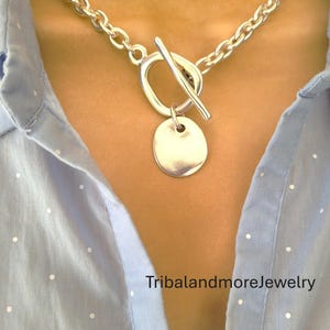 May include: A silver-toned necklace with a toggle clasp and a circular pendant. The chain is made of thick, rounded links. The necklace is worn over a light blue button-down shirt. The text "TribalandmoreJewelry" is visible.