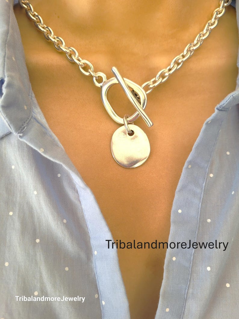 May include: A silver-toned necklace with a toggle clasp and a circular pendant. The chain is made of thick, rounded links. The necklace is worn over a light blue button-down shirt. The text "TribalandmoreJewelry" is visible.
