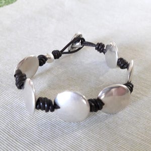 Puede incluir: Un brazalete de cuero negro con discos de metal plateado. El brazalete tiene un dise&ntilde;o sencillo y es ajustable.