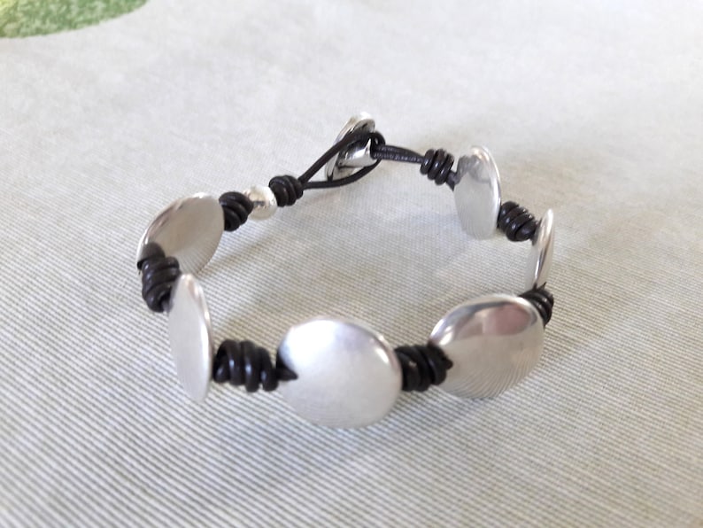 Puede incluir: Un brazalete de cuero negro con discos de metal plateado. El brazalete tiene un dise&ntilde;o sencillo y es ajustable.