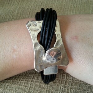 May include: A black leather bracelet with a silver, hammered, rectangular clasp. The clasp has a round, silver accent.