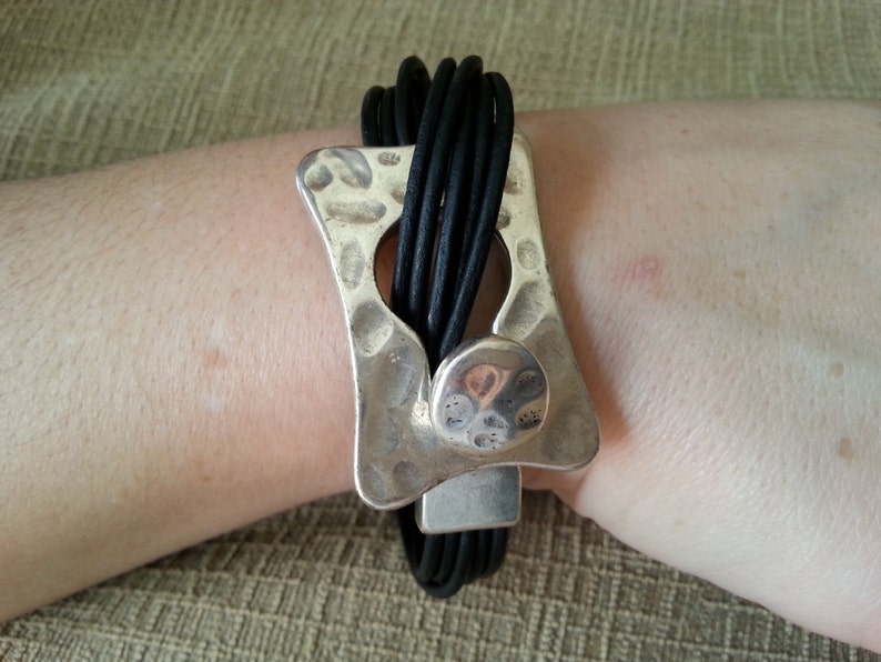 May include: A black leather bracelet with a silver, hammered, rectangular clasp. The clasp has a round, silver accent.