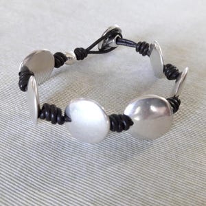 Puede incluir: Un brazalete de cuero negro con abalorios redondos de metal plateado. El brazalete tiene un dise&ntilde;o sencillo y es ajustable.