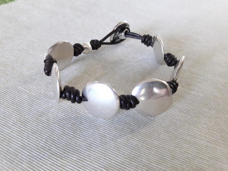 Puede incluir: Un brazalete de cuero negro con abalorios redondos de metal plateado. El brazalete tiene un dise&ntilde;o sencillo y es ajustable.
