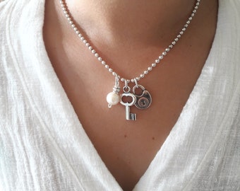 May include: Silver-toned necklace with a ball chain and three charms: a key, a padlock, and a pearl with a silver wire wrap. The necklace is worn against a white shirt.