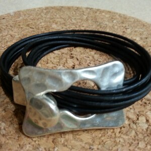 May include: A black leather bracelet with a silver, hammered metal clasp. The bracelet is wrapped around itself multiple times.
