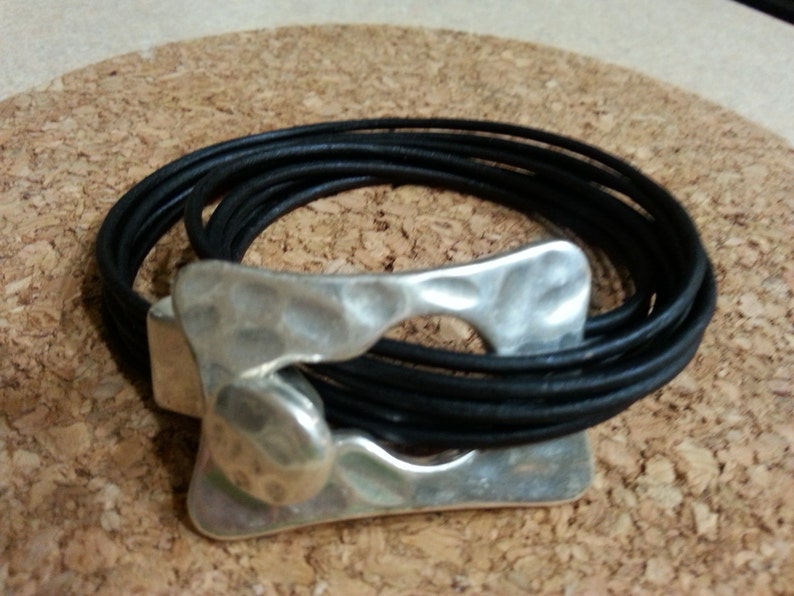 May include: A black leather bracelet with a silver, hammered metal clasp. The bracelet is wrapped around itself multiple times.