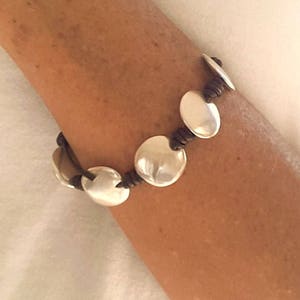 Puede incluir: Un brazalete de cuero marr&oacute;n con abalorios de disco de plata. El brazalete se lleva en la mu&ntilde;eca.