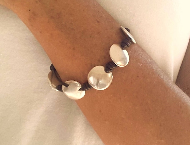 Puede incluir: Un brazalete de cuero marr&oacute;n con abalorios de disco de plata. El brazalete se lleva en la mu&ntilde;eca.