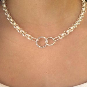 Peut inclure: Un collier argenté avec une chaîne épaisse. La chaîne est composée de maillons arrondis. Deux anneaux entrelacés forment le pendentif central. Le collier est porté autour du cou.