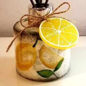 May include: A glass diffuser bottle with a silver lid and a lemon slice on top. The bottle has a white background with a lemon print and green leaves. Brown sticks are inserted into the bottle.