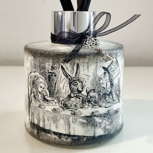 May include: A black and white diffuser bottle featuring an image of Alice in Wonderland characters enjoying tea. The bottle has a silver lid and a black ribbon with a silver key charm.