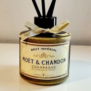 May include: A gold bottle with a black lid and a gold ribbon. The bottle has a label that says "Brut Impérial Moët & Chandon Champagne". The bottle is filled with black diffuser sticks.