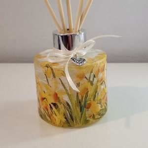 Può includere: Una bottiglia di diffusore in vetro con un tappo argentato e un nastro bianco. La bottiglia è decorata con un motivo floreale di narcisi gialli e foglie verdi. La bottiglia ha un ciondolo argentato a forma di cuore.