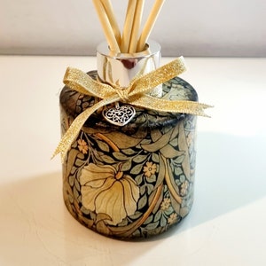 May include: A decorative reed diffuser with a floral pattern in shades of green, beige, and gold. The glass bottle has a silver collar, a gold ribbon, and a heart-shaped charm. Several wooden reeds are inserted into the bottle.