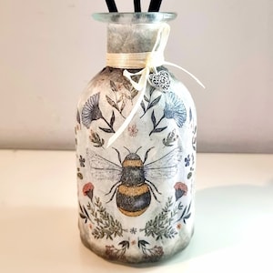 May include: A white glass diffuser bottle with a bee and floral design. The bottle has a cream-coloured ribbon tied around the neck and a silver charm in the shape of a heart. Four black diffuser sticks are in the bottle.