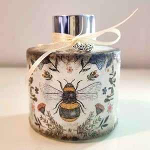 May include: A white ceramic diffuser bottle with a silver lid and a beige ribbon tied around the neck. The bottle has a floral and bee design with a small silver heart charm.