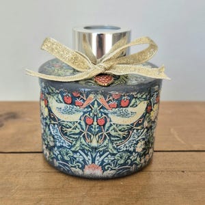May include: A decorative diffuser with a silver top and a gold ribbon. The cylindrical body features a pattern of birds, strawberries, and foliage in blue, green, and red. The diffuser is presented on a wooden surface.