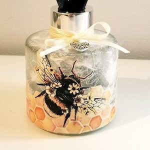 Peut inclure: Une bouteille de diffuseur avec une surface textur&eacute;e grise et blanche. La bouteille repr&eacute;sente une abeille avec du miel et des fleurs. La bouteille a un couvercle argent&eacute; et des b&acirc;tonnets diffuseurs noirs.