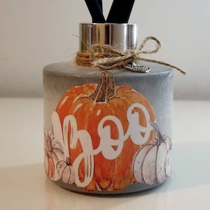 Może przedstawiać: Szary betonowy dyfuzor z czarnymi patyczkami. Butelka jest ozdobiona grafiką dyni i napisem "Boo". Srebrny kołnierz i kokarda ze sznurka dopełniają projekt.