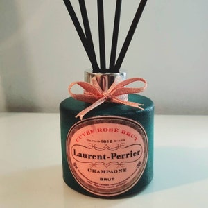 May include: A green glass bottle with a pink label that reads "Cuvée Rose Brut" and "Laurent-Perrier Champagne Brut" with a pink bow and black diffuser sticks.