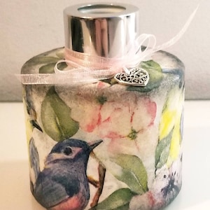 May include: A round glass diffuser bottle with a silver lid and a pink ribbon. The bottle is decorated with a floral pattern and a small bird. A silver heart charm hangs from the ribbon.