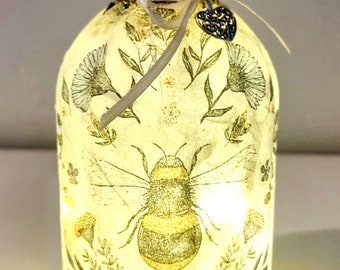 May include: A decorative glass bottle with a bee and floral design. The bottle is illuminated from within, creating a warm glow. A ribbon and heart-shaped charm adorn the neck of the bottle.
