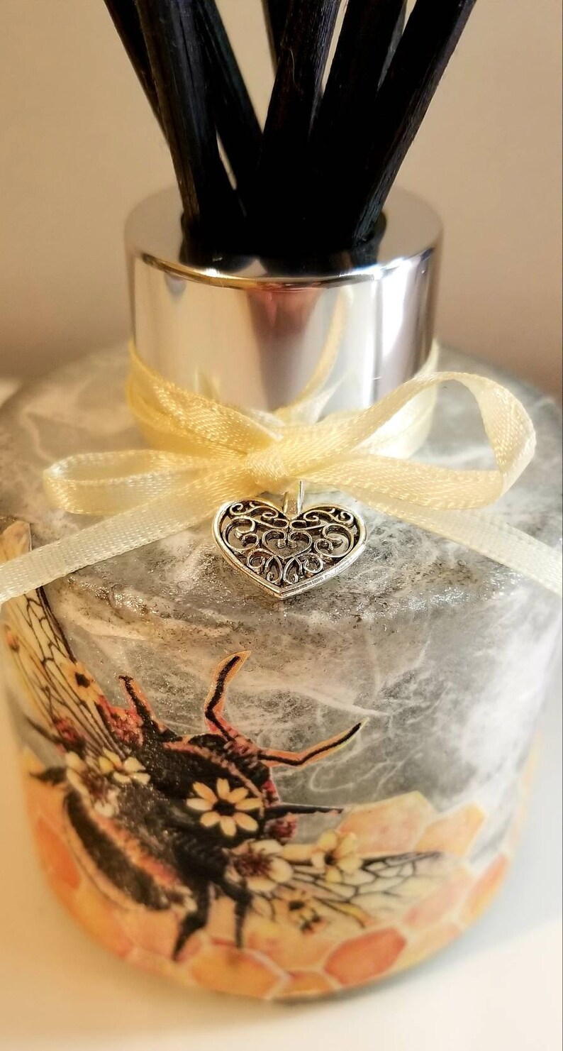 Peut inclure: Une bouteille de diffuseur en verre avec un motif d'abeille gris et orange. La bouteille a un couvercle argent&eacute; et un ruban jaune avec un charme de c&oelig;ur argent&eacute;.